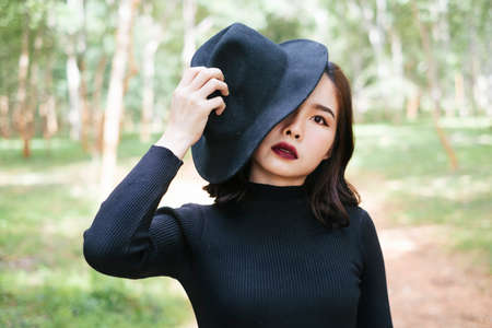 Woman in black dress cover half of her face with black hat, standing in the forrest.