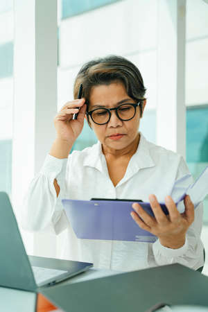Busy old elderly senior woman wearing eyeglasses at working desk in office room.