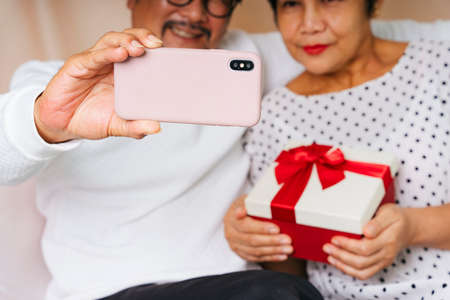 Asian senior couple taking selfie with smartphone in birthday party. Wife holding present gift box.の写真素材