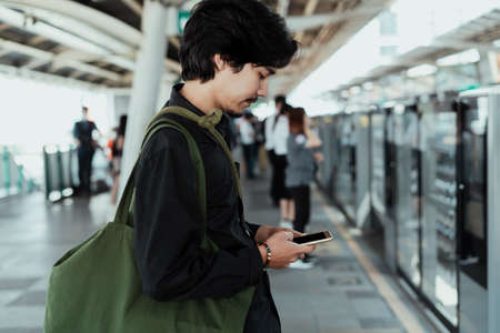 Black hair guy standing calmly at the skytrain station and use his mobile phone while waiting for the train.の写真素材