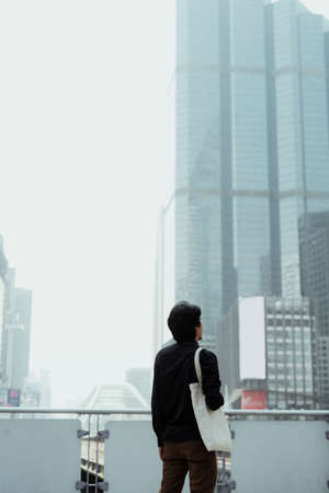 Black hair guy with white canvas bag standing at the skytrain station's terrace look at the high building in the city, vertical.の写真素材