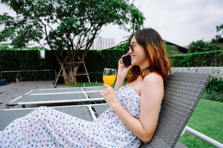 Beautiful asian woman wear eyeglasses using smartphone and hold orange glass on beach chair in hotel.の写真素材