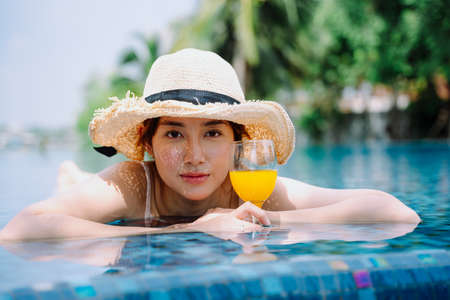 Beautiful asian woman traveler wear hat hold glass of orange juice relax in swimming pool.の写真素材