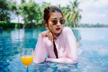 Beautiful asian woman traveler wear swim suit and sunglasses enjoy orange juice in swimming pool.