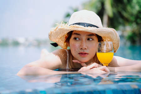 Beautiful asian woman traveler wear hat hold glass of orange juice relax in swimming pool.の写真素材