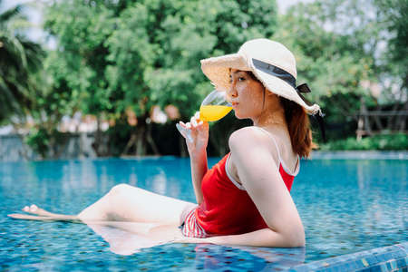 Side view - Beautiful asian woman traveler in red swim suit and hat drink orange juice on swimming pool.の写真素材