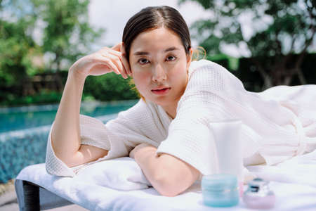 Portrait of asian woman in white bathrobe at spa treament bed with lotion bottle near swimming pool.