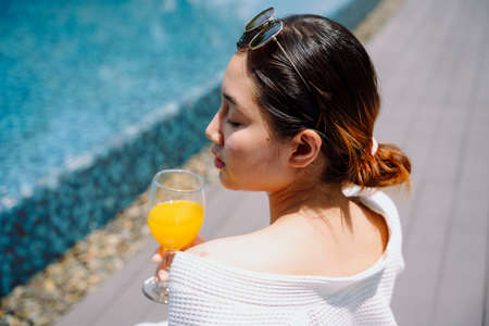 Back view - Beautiful asian woman traveler in white bathrobe enjoy drinking orange juice at poolside...
