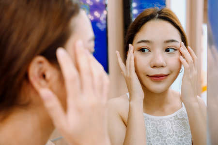 Asian woman looking at mirror checking her face in mirror after facial treatment at beauty clinic.の写真素材