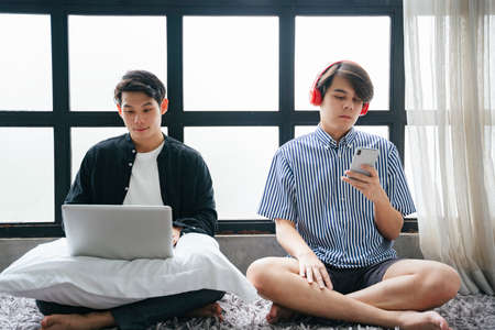 Couple of asian LGBTQ+ men living in the house, working on digital laptop computer and listening...
