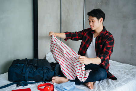 Asian young man packing clothes and stuff in luggage for a trip.