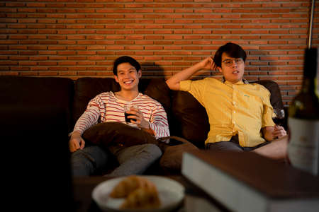 Couple of asian LGBTQ+ men enjoy wine and watch TV program together at home.