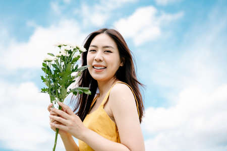 Portrait of beautiful asian woman in yellow dress hold white flowers on summer cloud background.