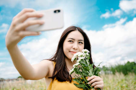 Beautiful asian woman hold white flowers taking selfie with smartphone on a field.の写真素材