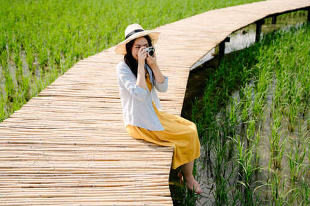 Beautiful asian traveler woman in yellow dress hold camera sitting on wooden bridge in rice field.