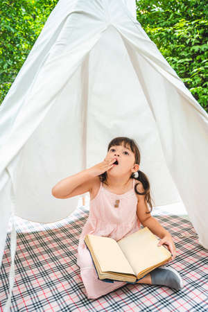 Cute asian little girl with a book sitting inside white tent at garden outdoors. She is looking up the sky with excited expression.の写真素材