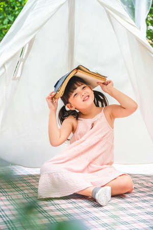 Cute asian little girl with a book covering head stay inside white camping tent at garden.の写真素材