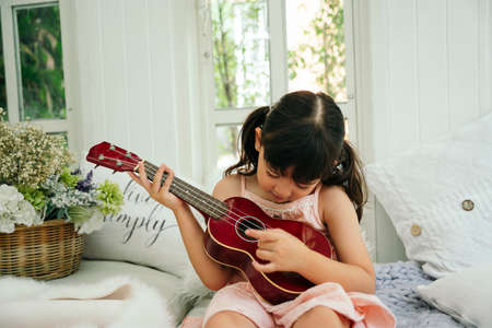 Asian child girl with wooden classic ukulele on sofa at home.