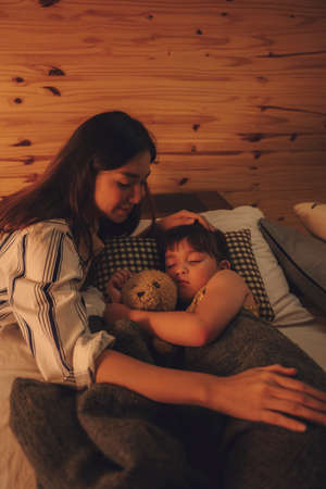 Asian mother watching her child sleep and hug teddy bear on a bed at night.の写真素材