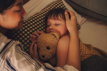 Asian mother watching her child sleep and hug teddy bear on a bed at night.の写真素材