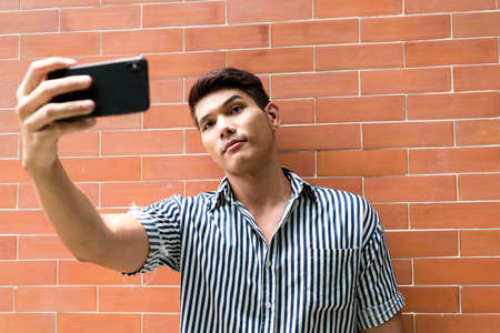 Asian man taking selfie with smartphone on brick wall background.の写真素材