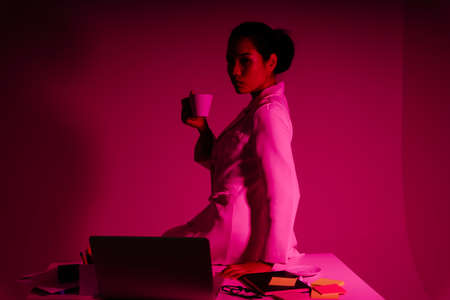 Portrait of businesswoman holding cup of coffee sitting on office desk in red neon light.