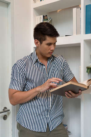 Asian man reading a book leaning on bookshelf in library.