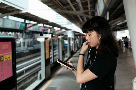 Young asian thai woman listen to music with earphones from smartphone, waiting for a train on train platform.の写真素材