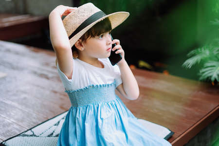 Little girl in white blue dress wearing hat talking on the phone at home.の写真素材