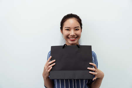 Cheerful and playful asian woman holding black shopping paper bag with two hands isolated on white background - Copy space.の写真素材