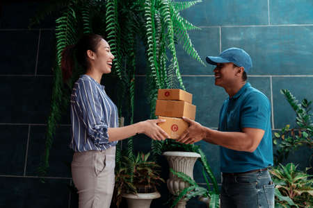 Smiling Asian courier man talking to customer giving cardboard boxes to cheerful woman.の写真素材