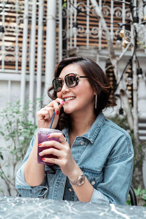 Asian thai woman on jeans jacket wearing sunglasses drinking grape juice from plastic cup with straw at cafe.の写真素材