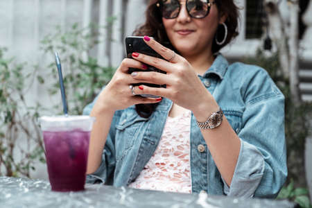 Cool Asian thai woman on jeans jacket wearing sunglasses playing smartphone at cafe. Grape soda in plastic cup on foreground.の写真素材