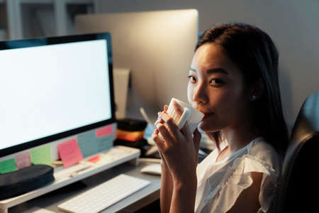 Asian thai woman drinking and sipping hot coffee from a cup, sitting infront of computer at office. Night shift time.の写真素材
