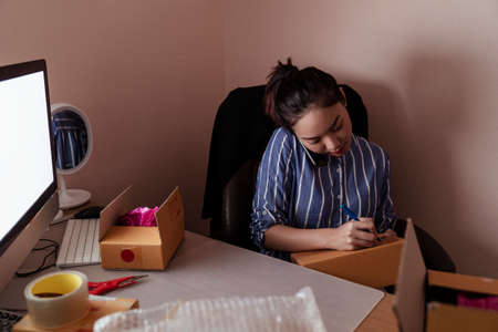 Busy asian thai woman writting address on cardboard box and talking on the phone with customer. Copy space on computer.の写真素材