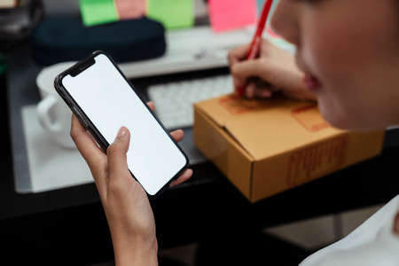 Hand of asian delivery woman reading information on smartphone then writing address down on cardboard box with red pen.の写真素材