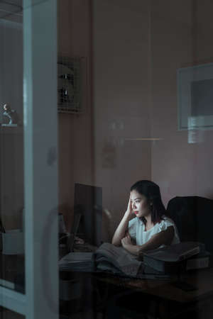 Stressful asian woman sitting in office room working on computer until midnight - working overtime concept.の写真素材