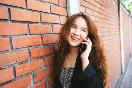 Fashion portrait of young asian woman wearing black jacket talking on the phone near brick wall.の写真素材