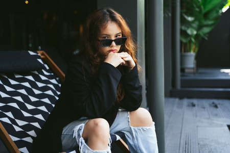 Fashion portrait of young asian woman wearing sunglasses and black jacket sitting on chair beach.