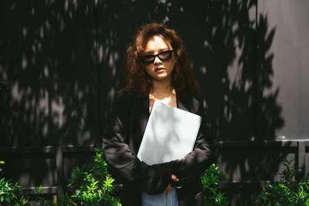 Fashion portrait of young asian woman wearing sunglasses and black jacket holding laptop computer.の写真素材