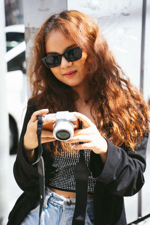 Fashion portrait of young asian woman wearing sunglasses and black jacket holding camera.