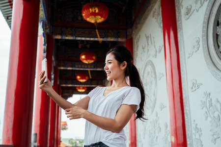 Cute asian woman on white shirt taking selfie with smartphone with right hand at Chinese temple.