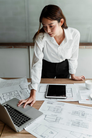 Busy long hair woman architect working hard with her laptop, lots of blueprint is on her table.の写真素材