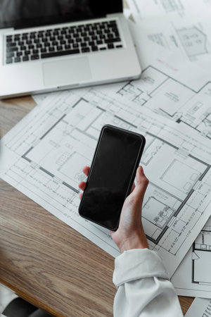 Cropped image of lots of blueprint on the wooden table, laptop and smartphone in a woman interior designer's hand while she searching the infoemation on the internet.の写真素材