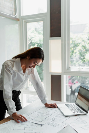 Long hair designer woman in white shirt standing at her wooden table look at the building plan in her hands.の写真素材