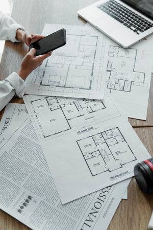 Cropped image of lots of blueprint on the wooden table, laptop and smartphone in a woman interior designer's hand while she searching the infoemation on the internet.の写真素材