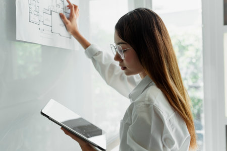 Long hair woman interior designer in white shirt wearing eyeglasses compare the blueprint on whiteboard with the blueprint in tablet.の写真素材