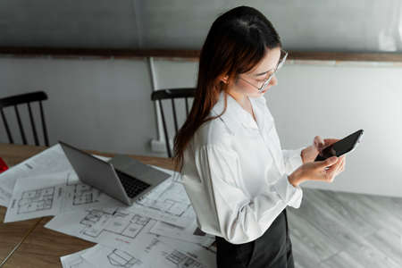 Long hair woman interior designer in white shirt wearing eyeglasses leaning at the wooden table full of paper reading the client's text in smartphone.の写真素材