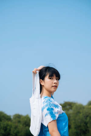 Black hair French braid girl in white and blue tie dye hold canvas bag at her back under clear sky,...