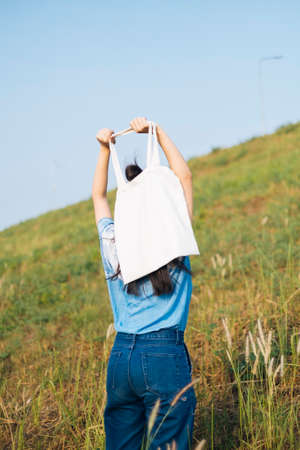 Back shot of black half tied hair girl in white and blue tie dye standing in slope grassland and raise the canvas bag up.の写真素材